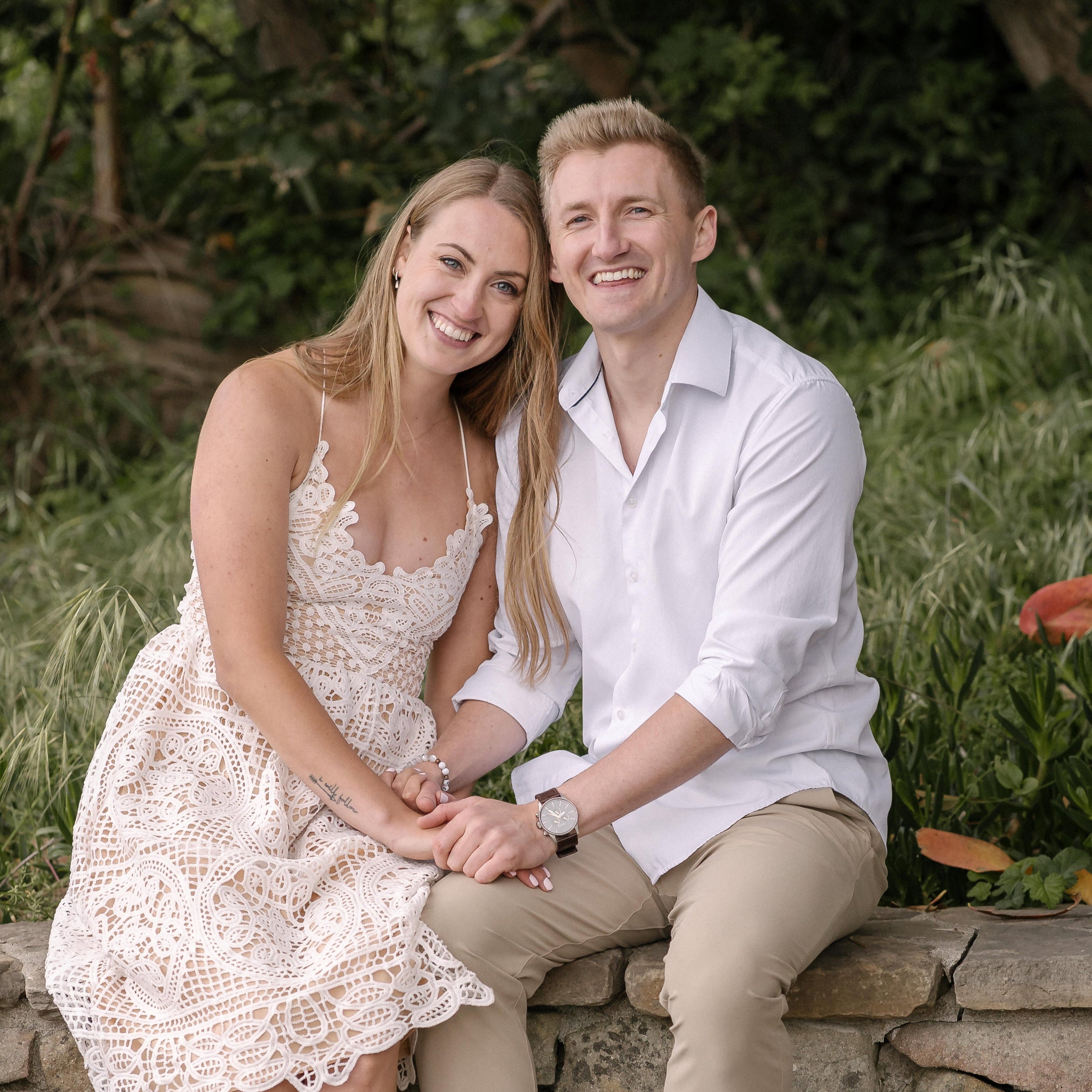 Couple sitting on a stone wall outdoors with greenery in the background